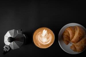 Latté Kunst Kaffee im ein Weiß Tasse zwei Seiten mit Italienisch Moka Töpfe, Kaffee und Croissants, oben Sicht, schwarz Hintergrund, leeren Raum, und drei Kreuzungen zum verwenden. foto