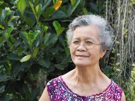 ein Senior asiatisch Frau mit kurz Weiß Haar lächelnd und suchen beim das Kamera während Stehen im ein Garten foto