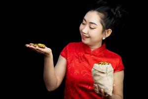 Porträt asiatische junge Frau rotes Kleid traditionelles Cheongsam hält eine Goldmünze in einem Sack auf dem schwarzen Hintergrund foto