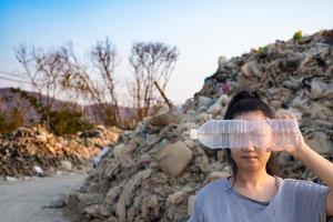 Frauenhand, die eine durchsichtige Plastikflasche hält, um das Gesicht am großen Müllhintergrund des Berges zu bedecken foto