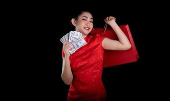 Porträt einer schönen jungen asiatischen Frau, rotes Kleid, traditionelles Cheongsam, das eine Tasche zum Einkaufen und Geldbanknote 100 usd auf schwarzem Hintergrund hält