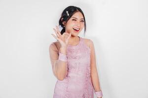 attraktiv jung asiatisch Frau im Rosa Kleid ist gestikulieren in Ordnung Zeichen mit ihr Finger, isoliert durch Weiß Hintergrund foto