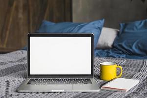 Laptop leerer weißer Bildschirm in der Nähe von Tasse Spiralnotizblock Bett