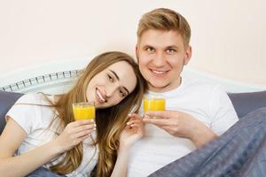 glückliches Paar, das morgens Orangensaft trinkt, während es im Bett im Schlafzimmer liegt - gesunder Lebensstil und Ernährungskonzept