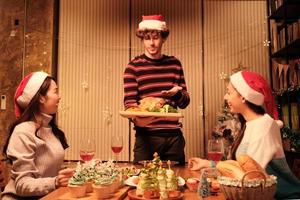 Das besondere Essen der Familie, junger Mann serviert gebratenen Truthahn an Freunde, fröhlich mit Getränken und genießt das Essen, Abendessen im Esszimmer des Hauses, das für Weihnachtsfeste und Neujahrsfeiern dekoriert ist. foto