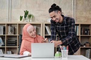 Zwei junge, islamisch geprägte Startup-Kollegen erzählen mit einem Lächeln über die erfolgreiche Arbeit in einem E-Commerce-Unternehmen. Laptop zur Online-Kommunikation über das Internet in einem kleinen Büro verwenden.