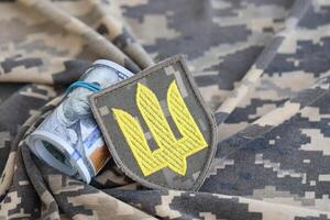 ukrainisch Heer Symbol und Bündel von Dollar Rechnungen auf Militär- Uniform. Zahlungen zu Soldaten von das ukrainisch Heer von vereinigt Zustände, Gehälter zu das Militär. Krieg Unterstützung foto