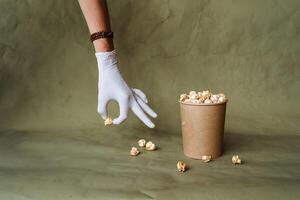 Hand im ein Weiß Handschuh erreicht zum Popcorn, nehmen Essen im schützend Handschuhe, halt ein Stück von Essen mit zwei Finger, ein voll Box von Popcorn, Sicherheit beim das schnell Essen Verteilung, minimal Konzept foto