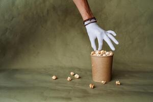 Hand im ein Weiß Handschuh erreicht zum Popcorn, nehmen Essen im schützend Handschuhe, halt ein Stück von Essen mit zwei Finger, ein voll Box von Popcorn, Sicherheit beim das schnell Essen Verteilung, minimal Konzept foto