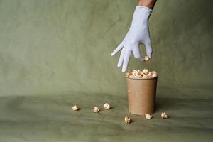 Hand im ein Weiß Handschuh erreicht zum Popcorn, nehmen Essen im schützend Handschuhe, halt ein Stück von Essen mit zwei Finger, ein voll Box von Popcorn, Sicherheit beim das schnell Essen Verteilung, minimal Konzept foto