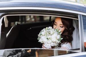 ein schön Braut, Sitzung im ein Auto, sieht aus aus von das öffnen Fenster von das Wagen. ein schön Braut mit ein Strauß von Blumen im ihr Hände ist Sitzung im ein stilvoll teuer Wagen. foto