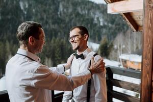 ein Vater Umarmungen seine reifen Sohn und hilft ihm bereiten zum das Hochzeit Zeremonie. warm und aufrichtig Beziehungen zwischen ein Vater und ein Erwachsene Sohn. ein emotional Moment beim ein Hochzeit foto