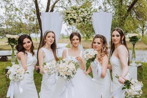 Gruppe Porträt von das Braut und Brautjungfern. Braut im ein Hochzeit Kleid und Brautjungfern im Weiß Kleider und halten stilvoll Blumensträuße auf das Hochzeit Tag gegen das Hintergrund von das See. foto