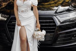 ein dunkelhaarig Braut steht Nächster zu ein modern schwarz Auto auf Ihre Hochzeit Tag mit ein Strauß von Blumen. Porträt von das Braut. Weiß Kleid, lange Schleier foto
