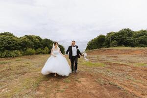 Porträt von ein jung Braut und Bräutigam Gehen auf Grün Gras gegen das Hintergrund von Berge nach das Hochzeit Zeremonie, Vorderseite Sicht. glücklich Hochzeit Paar, Kopieren Raum foto