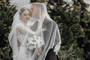 das Braut im ein elegant Kleid mit ein Strauß und das Bräutigam im ein klassisch passen sind Gehen im das Garten, halten Hände. foto