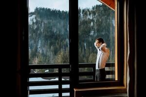Seite Aussicht von ein Mann im Hosenträger tragen ein Bogen Krawatte während Stehen auf ein Balkon mit Blick auf schneebedeckt Berge. sich ausruhen im das Berge. Berg Luft. Hochzeit im Winter foto