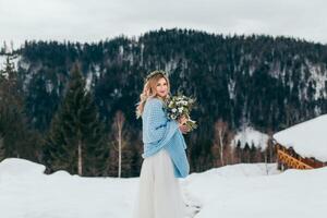 Porträt von ein süß Braut im ein Weiß Kleid mit ein Strauß von Blumen im ihr Hände, eingewickelt im ein Blau Plaid, warten zum das Bräutigam gegen das Hintergrund von ein Winter Landschaft von schneebedeckt Berge. foto