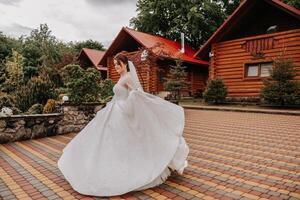 ein rothaarig Braut ist kreisen im ihr Hochzeit Kleid, gegen das Hintergrund von Berge und hölzern Häuser. großartig Kleid mit lange Ärmel, öffnen Büste. foto