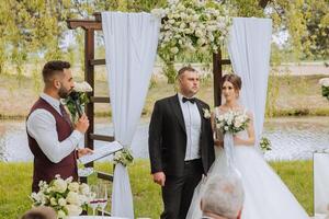 ein Bräutigam im ein schwarz passen und ein Braut im ein voluminös Kleid Stand in der Nähe von ein Weiß Bogen dekoriert mit Blumen während ein Hochzeit Zeremonie. das Braut ist halten ein Strauß. Frühling Hochzeit foto