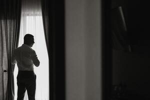 entspannt Mann setzt auf Weiß Hemd im Hotel Zimmer mit natürlich Licht. das Bräutigam trägt ein Hemd und bereitet vor zum das Hochzeit. kostenlos Raum foto