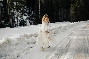 das Braut und Bräutigam sind Laufen entlang ein schneebedeckt Straße gegen das Hintergrund von ein Kiefer Wald und schön Kontrast Sonnenlicht. Seite Sicht. Winter Hochzeit. Platz zum Logo. foto