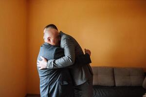 ein Vater Umarmungen seine reifen Sohn und hilft ihm bereiten zum das Hochzeit Zeremonie. warm und aufrichtig Beziehungen zwischen ein Vater und ein Erwachsene Sohn. ein emotional Moment beim ein Hochzeit foto