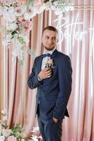 das Bräutigam im ein schwarz passen passt sich an seine Ansteckblume, posiert gegen das Hintergrund von ein Rosa Foto Verkaufsstand. Hochzeit Porträt.