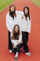 Porträt von drei Teenager Mädchen im beiläufig Kleider Sitzung im ein Stadion und posieren suchen beim das Kamera. Konzept von Freundschaft. ein Moment von Glück. foto