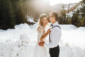 Porträt von das Braut und Bräutigam gegen das Hintergrund von ein Kiefer Wald und Hintergrundbeleuchtung. das Braut im ein Weiß Hochzeit Kleid, das Bräutigam im ein Weiß Hemd und Hose mit Hosenträger. Winter Hochzeit. foto