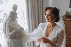 ein glücklich Braut ist vorbereiten zum ihr luxuriös Hochzeit im ein Hotel Zimmer, mit ein Hochzeit Kleid auf ein Mannequin nahe. Porträt von ein Frau mit modisch Haar, bilden und ein Lächeln im ein Dressing Kleid. foto