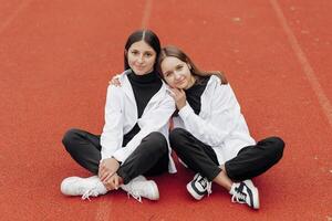 Porträt von zwei Teenager Mädchen im beiläufig Kleider Sitzung im ein Stadion und posieren suchen beim das Kamera. Konzept von Freundschaft. ein Moment von Glück. foto