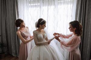 Morgen von das Braut. das Braut Maid von Ehre hilft das Braut Spitze oben ihr Kleid, befestigen Tasten auf das Kleid oder Ärmel. Freundinnen Hilfe das Braut befestigen ihr Kleid foto