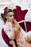 Porträt von ein jung schön elegant Frau Sitzung und posieren auf ein rot Wildleder- Sessel im Kunst Jugendstil Stil. schön Modell- Mädchen im ein luxuriös Abend Kleid. Dame mit perfekt bilden. foto