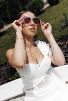 ein Mädchen im ein Weiß Kleid und Rosa Sonnenbrille im Natur. sich ausruhen im sonnig Wetter foto