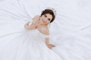 Brünette Braut im weg von der Schulter Spitze Weiß Kleid und Tiara posieren mit ein Strauß von Weiß und Rosa Blumen während Sitzung auf Grün Gras. schön Haar und bilden. Frühling Hochzeit foto