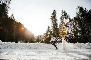 das Braut und Bräutigam sind Laufen entlang ein schneebedeckt Straße gegen das Hintergrund von ein Kiefer Wald und schön Kontrast Sonnenlicht. Seite Sicht. Winter Hochzeit. Platz zum Logo. foto