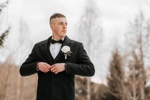 Hochzeit Porträt von das Bräutigam. das Bräutigam ist Stehen in der Nähe von ein Stein Zaun. ein Mann im ein schwarz Anzug. Winter Hochzeit foto