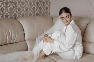 das Braut im ein Seide passen und Schleier posiert im ihr Zimmer, Sitzung auf das Couch. Morgen von das Braut. Vorbereitung. Hochzeit Fotografie foto