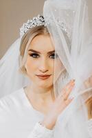 Hochzeit Porträt. ein blond Braut im ein Seide passen und Tiara posiert während Sitzung im ein Stuhl im ihr Zimmer foto