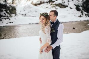 Porträt von das Braut und Bräutigam gegen das Hintergrund von ein Kiefer Wald und ein Fluss. das Braut im ein Weiß Hochzeit Kleid, das Bräutigam im ein Weiß Hemd und Hosenträger. Winter Hochzeit. foto