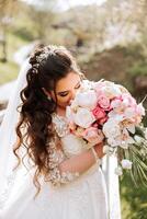 lockig Brünette Braut posiert mit ein Strauß, in der Nähe von ein hölzern Geländer. großartig Kleid mit lange Ärmel, öffnen Büste. Frühling Hochzeit foto