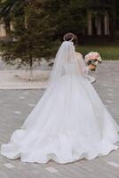 das Braut im ein großartig Kleid und Schleier Spaziergänge Nieder das Stein Pfad mit ihr Schultern gedreht. großartig Kleid mit lange Ärmel, öffnen Büste. Sommer- Hochzeit foto