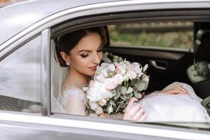 ein schön Braut, Sitzung im ein Auto, sieht aus aus von das öffnen Fenster von das Wagen. ein schön Braut mit ein Strauß von Blumen im ihr Hände ist Sitzung im ein stilvoll teuer Wagen. foto