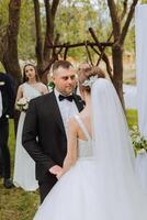 das Braut und Bräutigam halt Hände während das Gelübde in der Nähe von das Weiß Bogen dekoriert mit Blumen während das Hochzeit Zeremonie. das Braut ist halten ein Strauß. Frühling Hochzeit. glücklich Momente foto