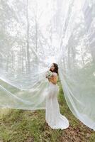 ein schön Braut ist Stehen im ein Grün Frühling Wald im ein glamourös Weiß Hochzeit Kleid mit ein Aussicht von das Schleier. ein romantisch Braut im ein Weiß Kleid ist posieren gegen das Hintergrund von das Wald. foto