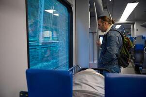 jung kaukasisch Mann Tourist mit Rucksack, Reiten ein Zug, Stehen beim Tür bereit zu aussteigen auf das Eisenbahn Bahnhof foto