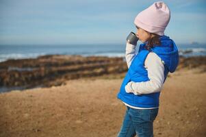 süß Kind Mädchen hält einer Hand im Tasche, Stehen auf das Strand und Trinken Tee von ein Thermosflasche Tasse, bewundern das Meer foto
