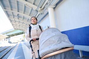 glücklich jung Frau schieben Baby Kinderwagen während Gehen auf das Plattform von ein Eisenbahn Bahnhof foto