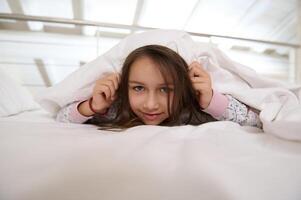kaukasisch boshaft Kind Mädchen im Pyjama, versteckt sich unter Weiß Decke während Lügen auf ihr Bett im Schlafgemach im das Morgen foto